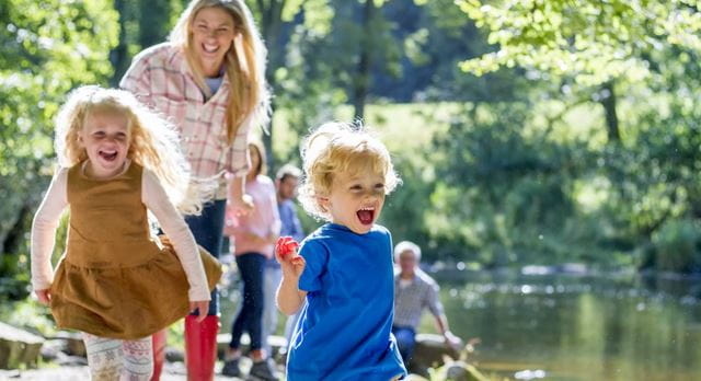 Two children running towards the camera with their mother trailing behind beside a river with the sun shining behind them